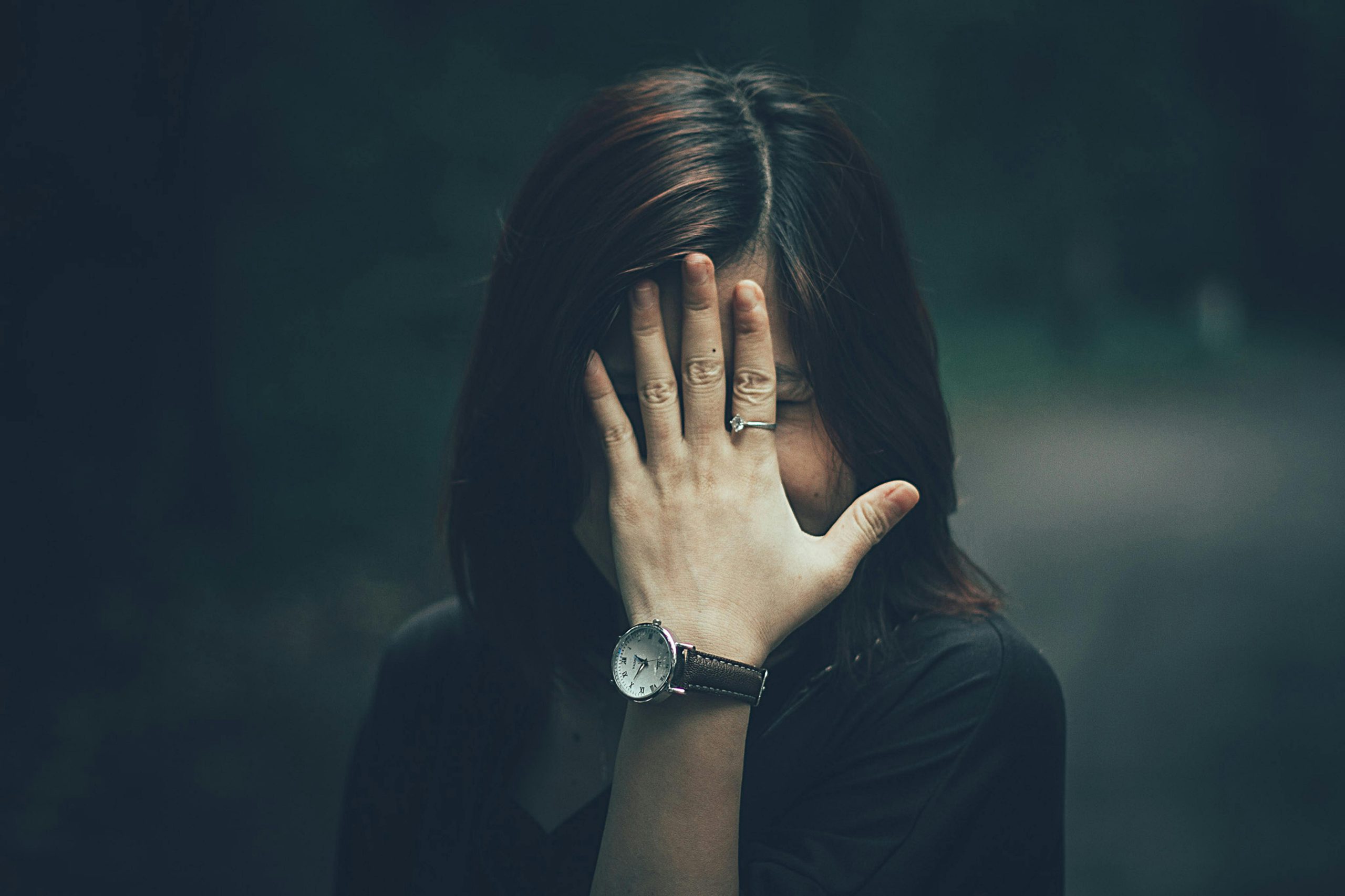 Une femme avec une montre à son poignet cache son visage avec sa main dans un cadre extérieur mélancolique.