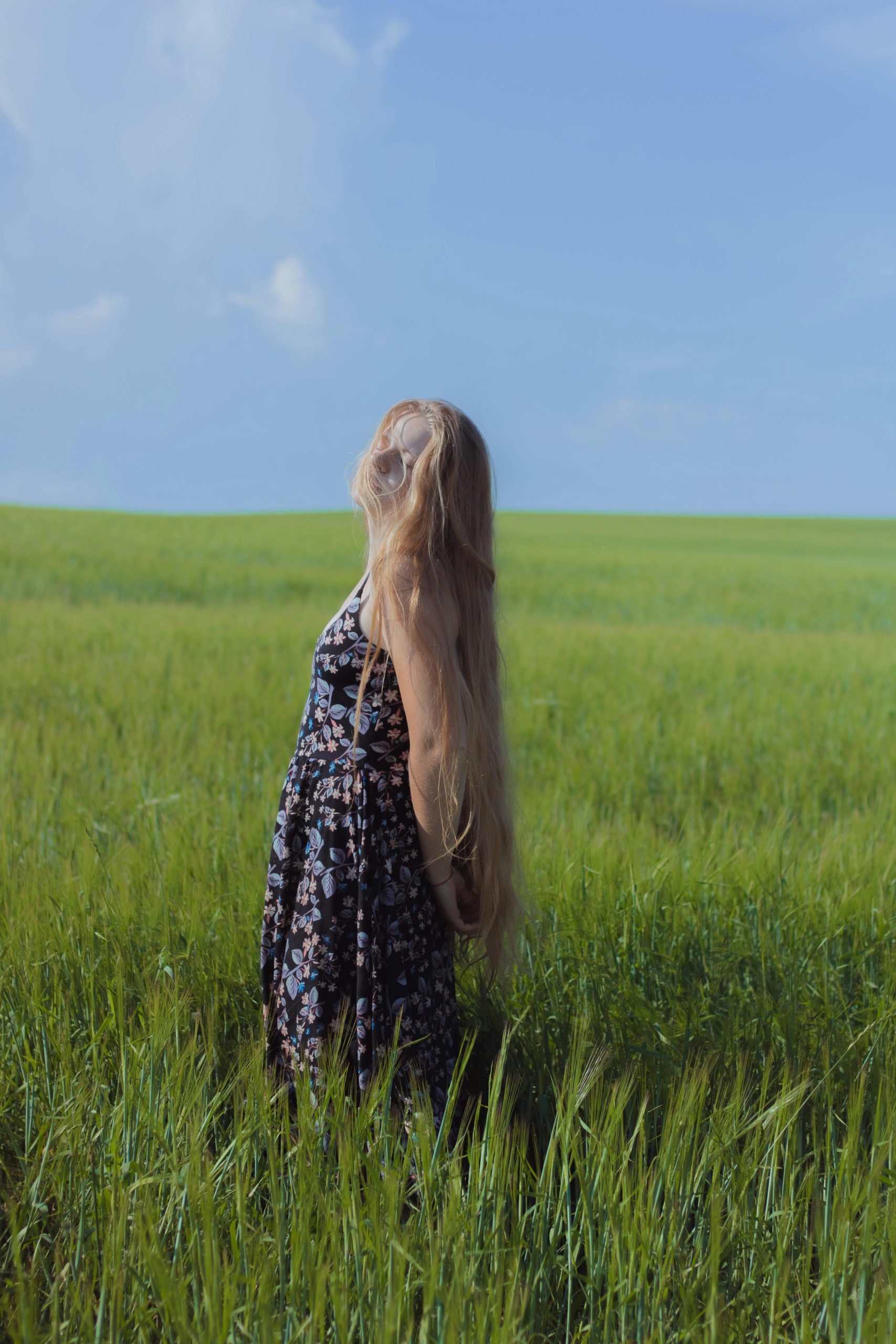 A blonde woman stands in a green field wearing a floral dress under a clear blue sky. Une femme blonde se tient dans un champ vert portant une robe fleurie sous un ciel bleu clair.