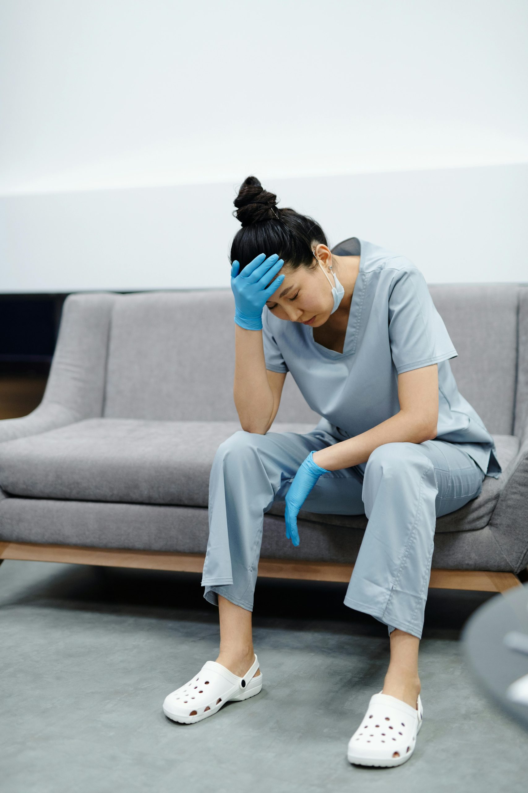 Tired nurse in scrubs sitting on a sofa, looking exhausted with her hand on her head. Infirmière fatiguée en uniforme assise sur un canapé, l'air épuisée avec sa main sur sa tête.