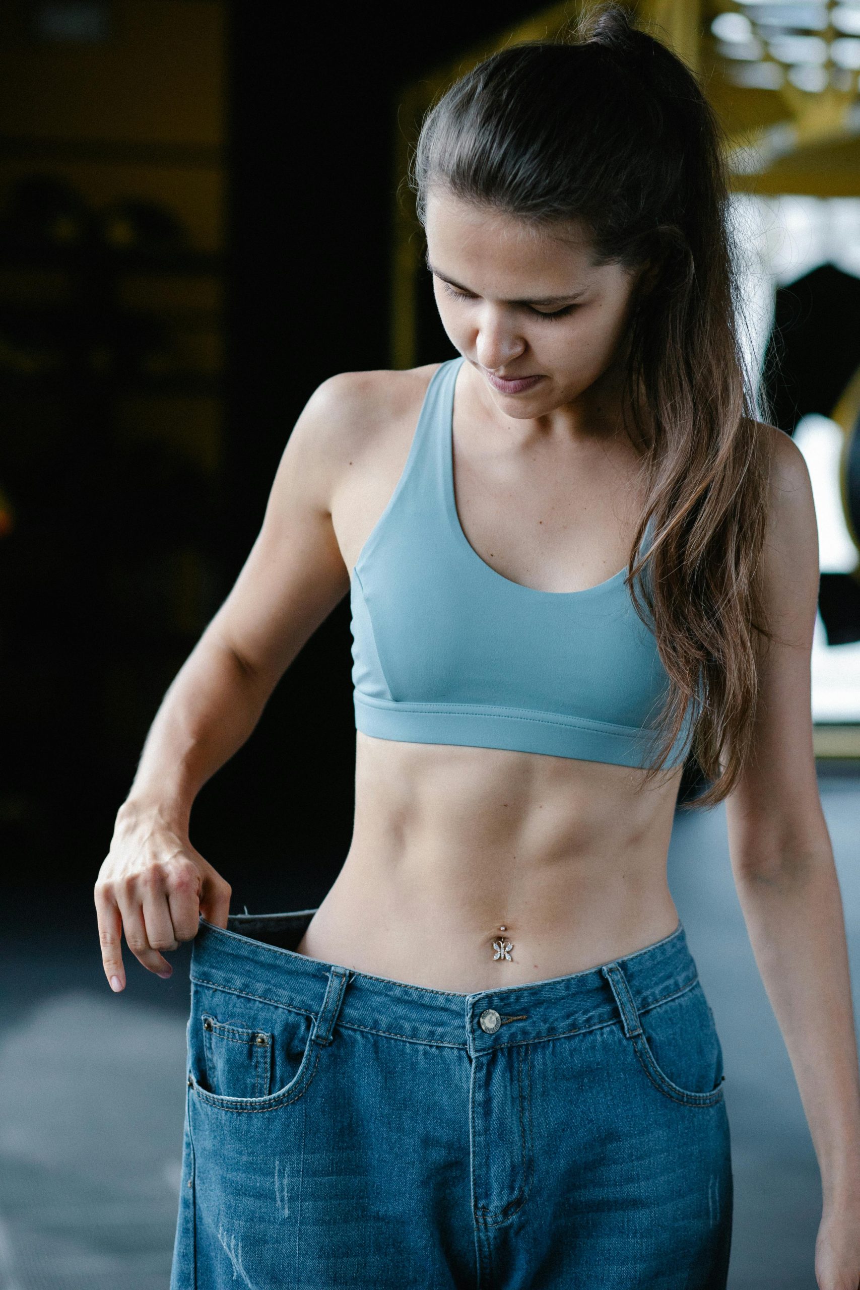 Fit woman showing weight loss success by wearing oversized jeans in gym. Femme en forme montrant son succès de perte de poids en portant un jean surdimensionné à la salle de sport.