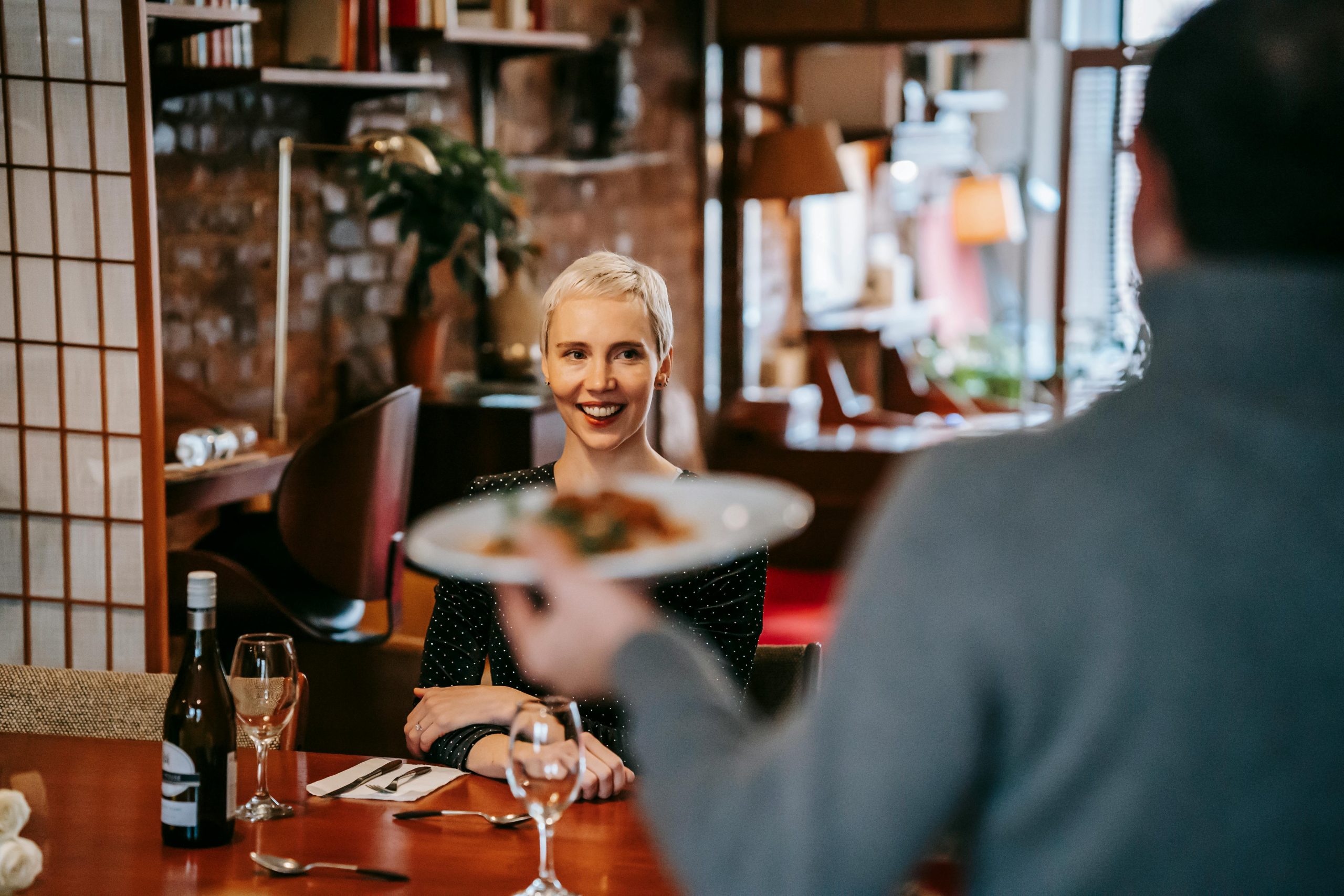 Couple positif ayant un rendez-vous dans un restaurant lumineux tandis qu'un homme servant une assiette de nourriture à une femme assise sur une chaise à une table avec une bouteille de vin et des verres près des couverts avec une serviette