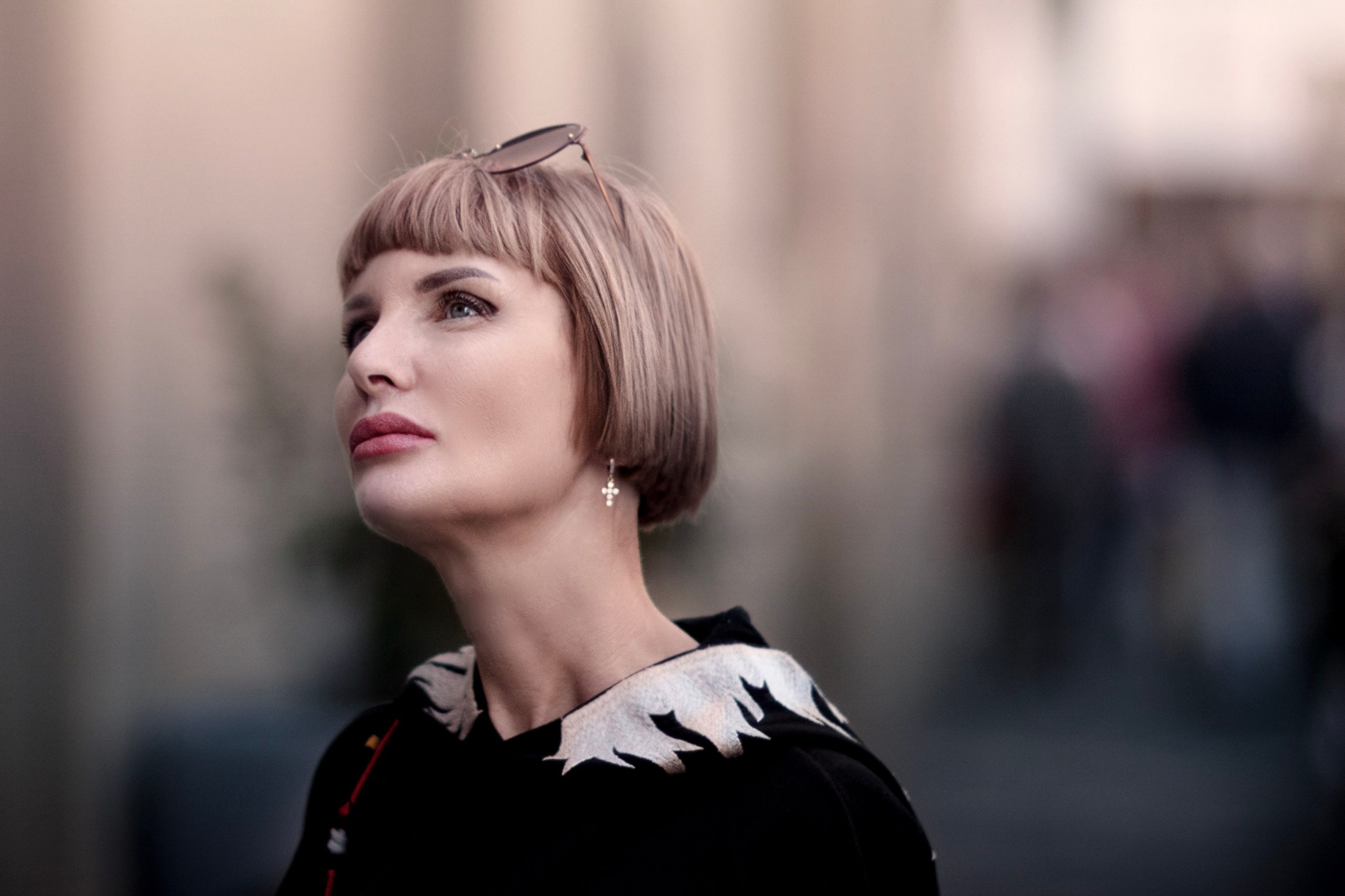 Stylish portrait of a woman with blonde bob haircut and sunglasses, looking up with a blurred urban background. Portrait élégant d'une femme avec une coupe bob blonde et des lunettes de soleil, regardant vers le haut avec un arrière-plan urbain flou.