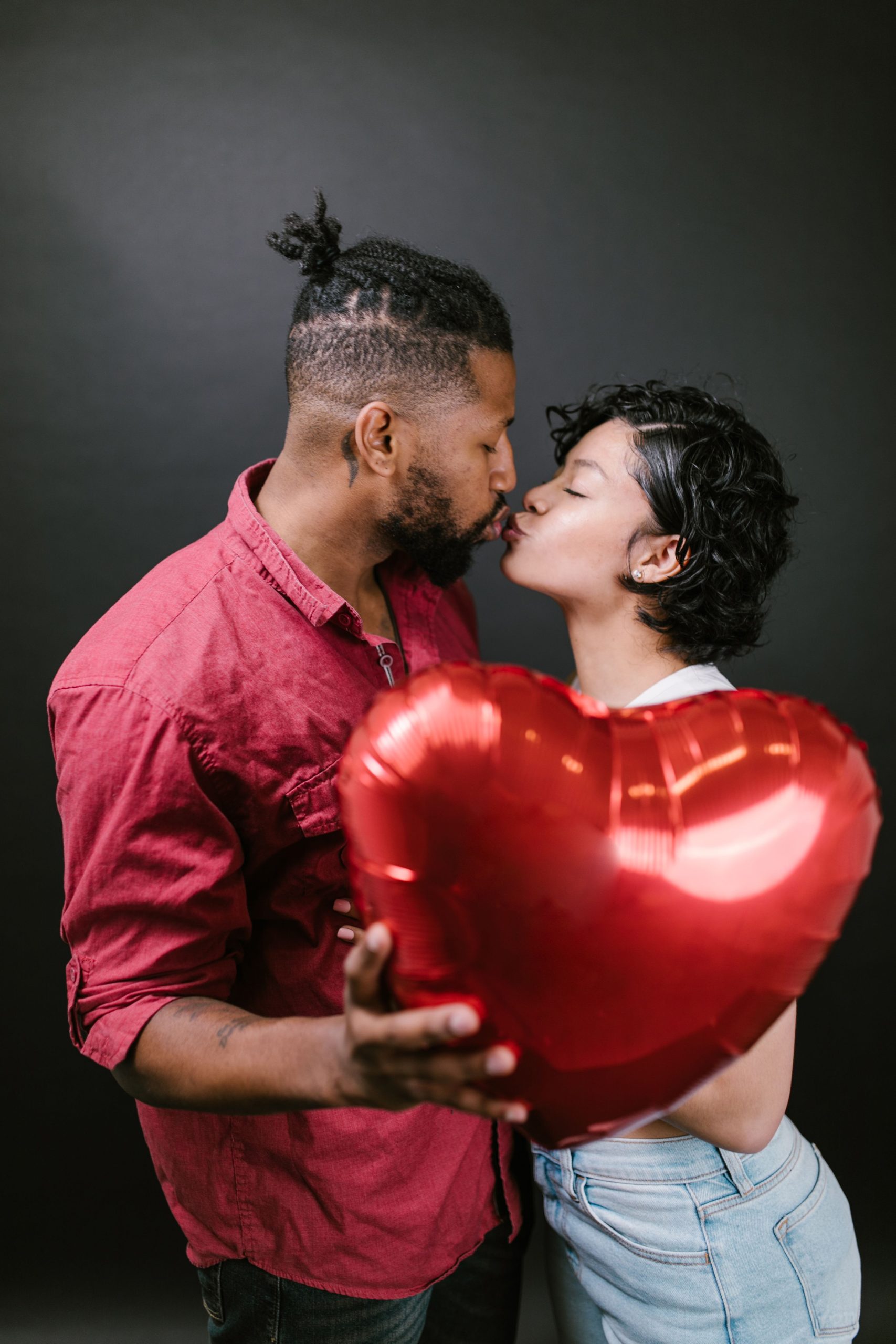 Couple kissing, holding a red heart balloon on a black background. Un couple s'embrasse en tenant un ballon en forme de cœur rouge sur un fond noir.