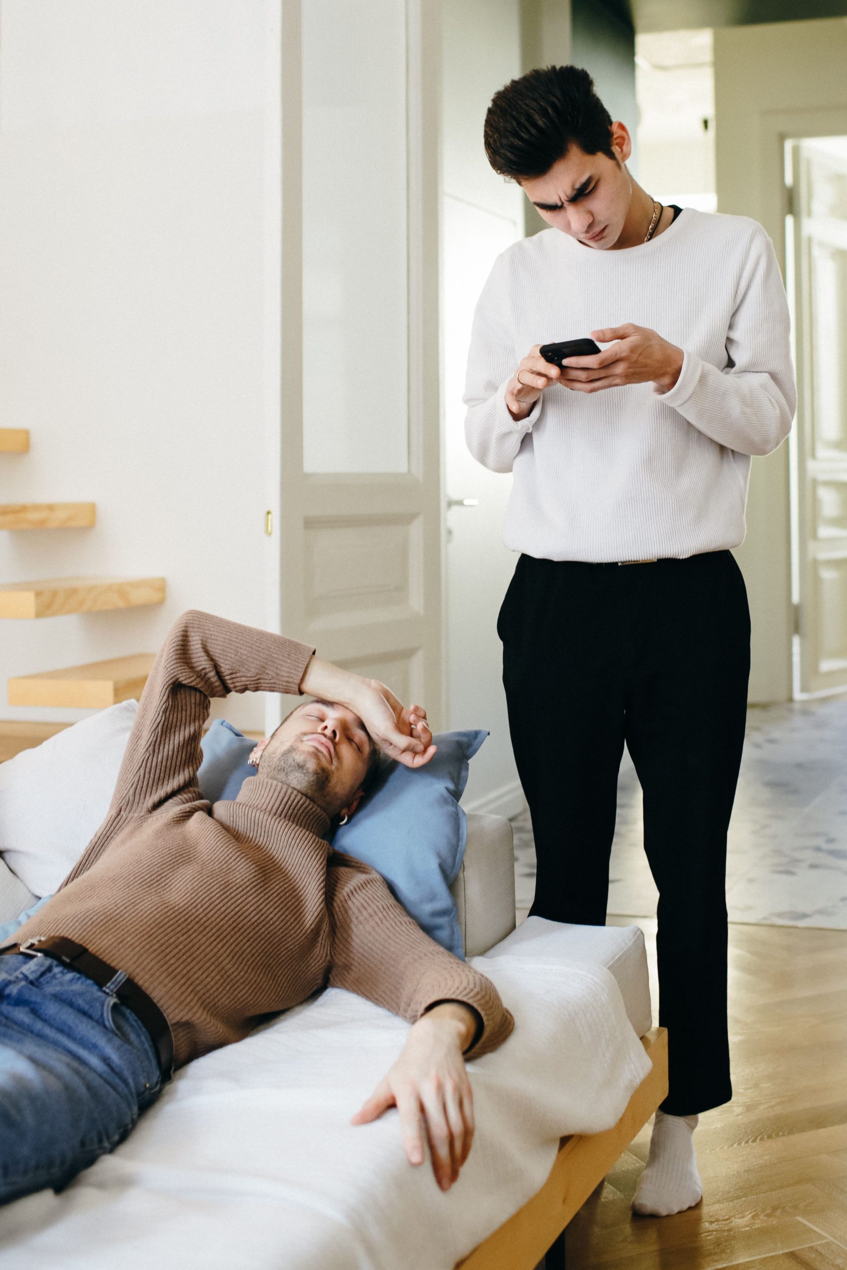 A young man in a white sweater checks his phone while a man in a brown sweater rests on the couch, portraying modern relationship dynamics. Un jeune homme en pull blanc vérifie son téléphone tandis qu'un homme en pull marron se repose sur le canapé, illustrant la dynamique relationnelle moderne.